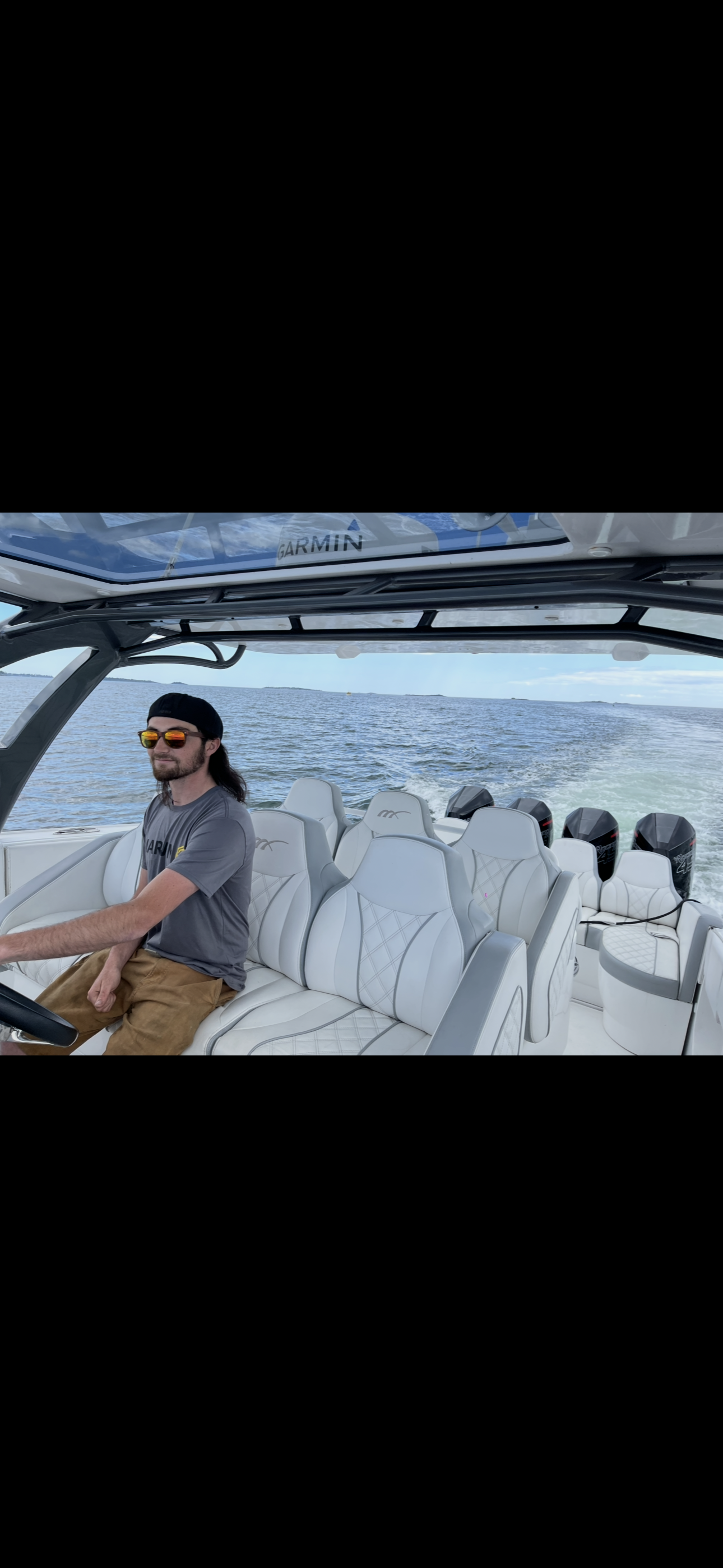 A person wearing sunglasses and a t-shirt driving a boat with white seats on open water under a bright sky.