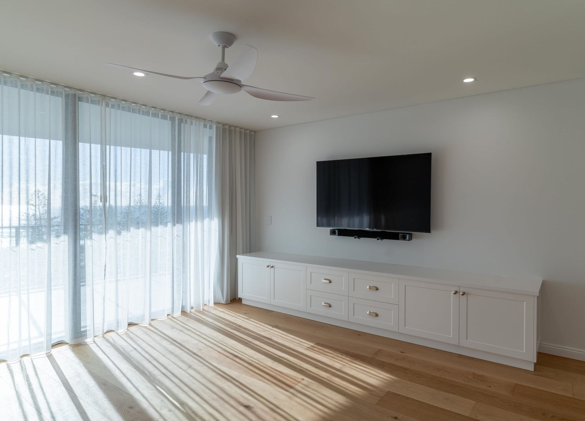 Bright living room with white cabinetry, a large TV, sheer curtains, and a ceiling fan.