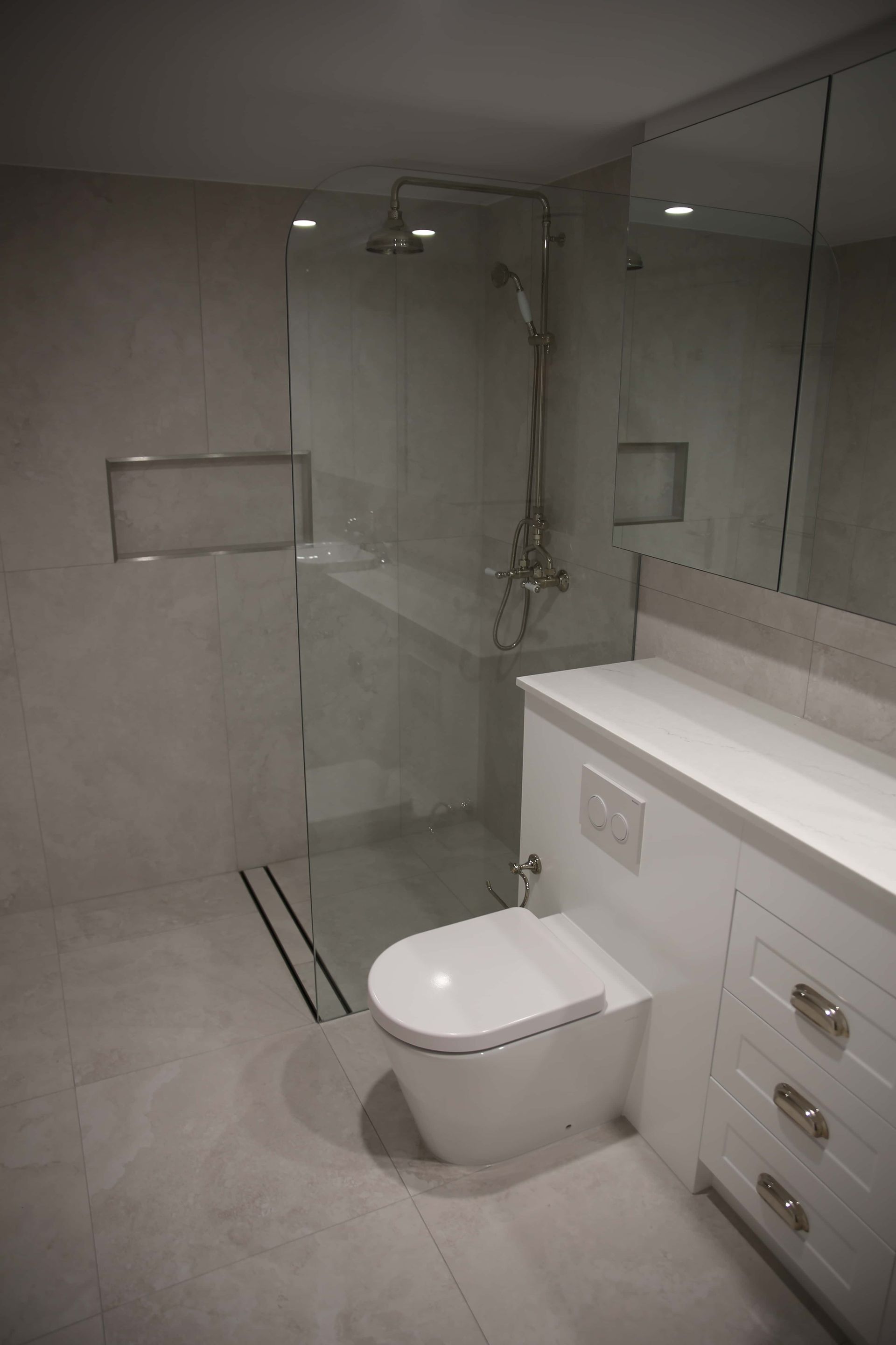 Modern white bathroom with walk-in shower, toilet, and vanity. Gray tile floor and walls; large mirror.