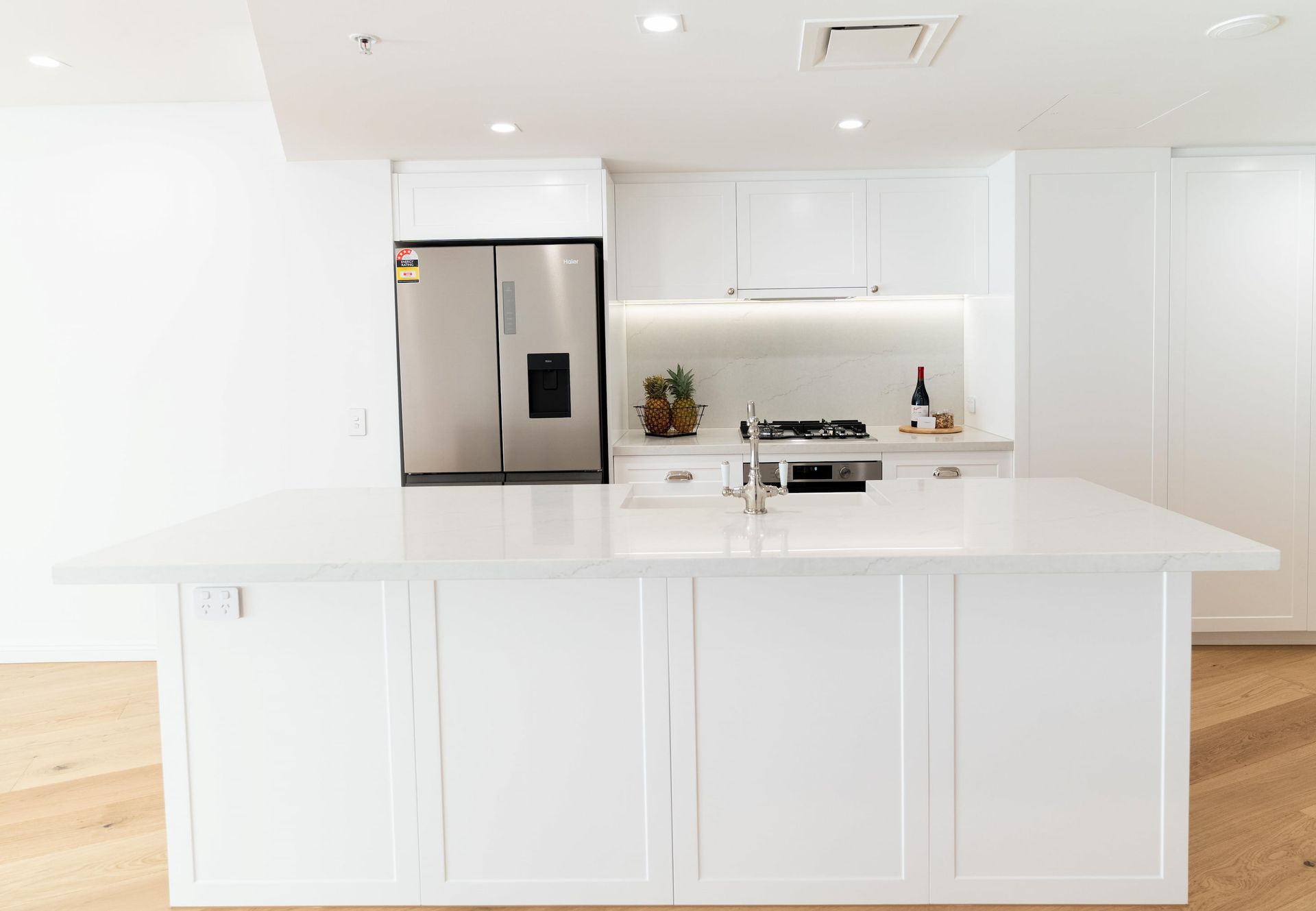 Modern white kitchen with island, stainless steel refrigerator, and light wood floors.