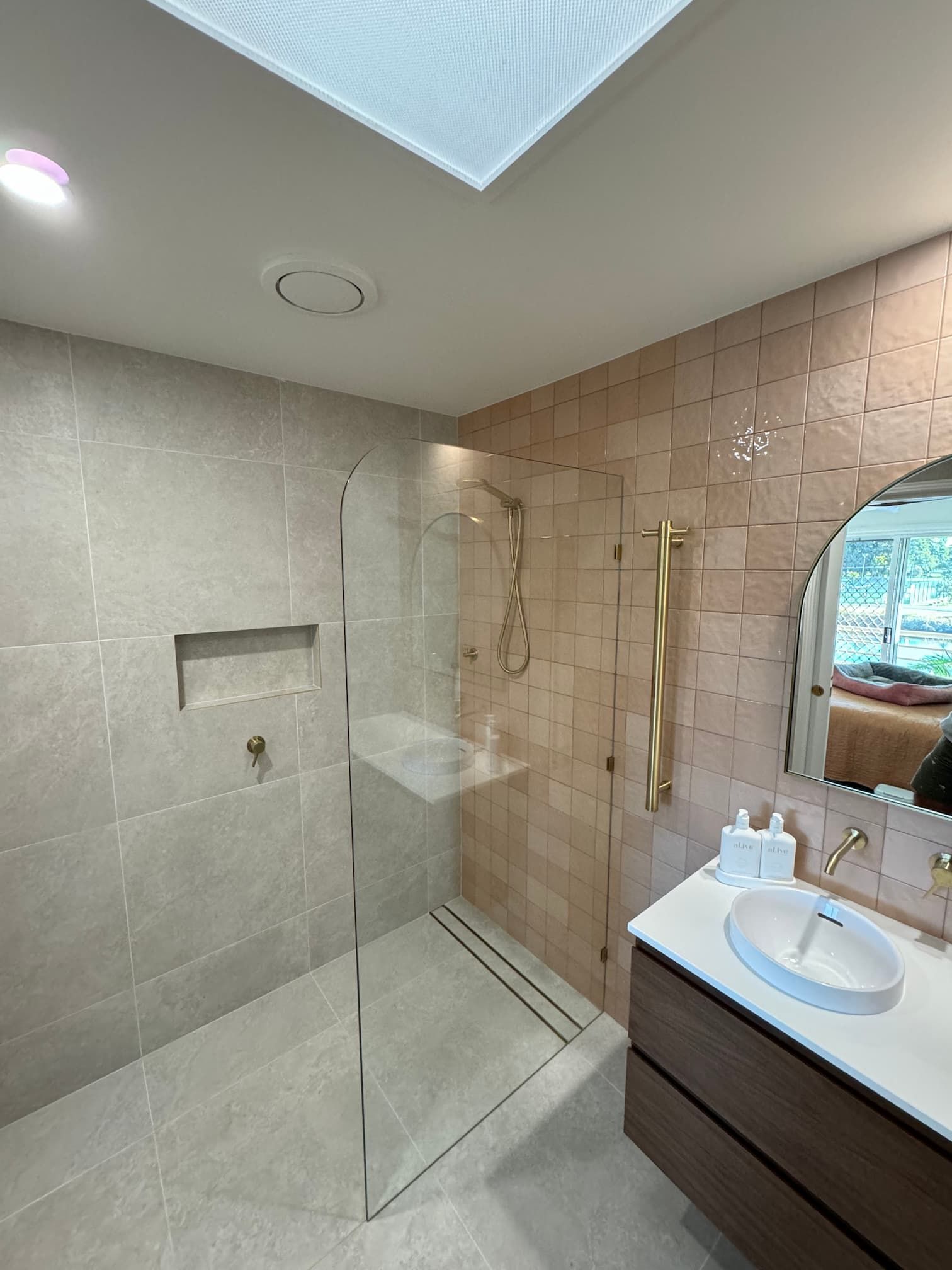 Modern bathroom with shower, vanity, and skylight; neutral tones with golden fixtures.