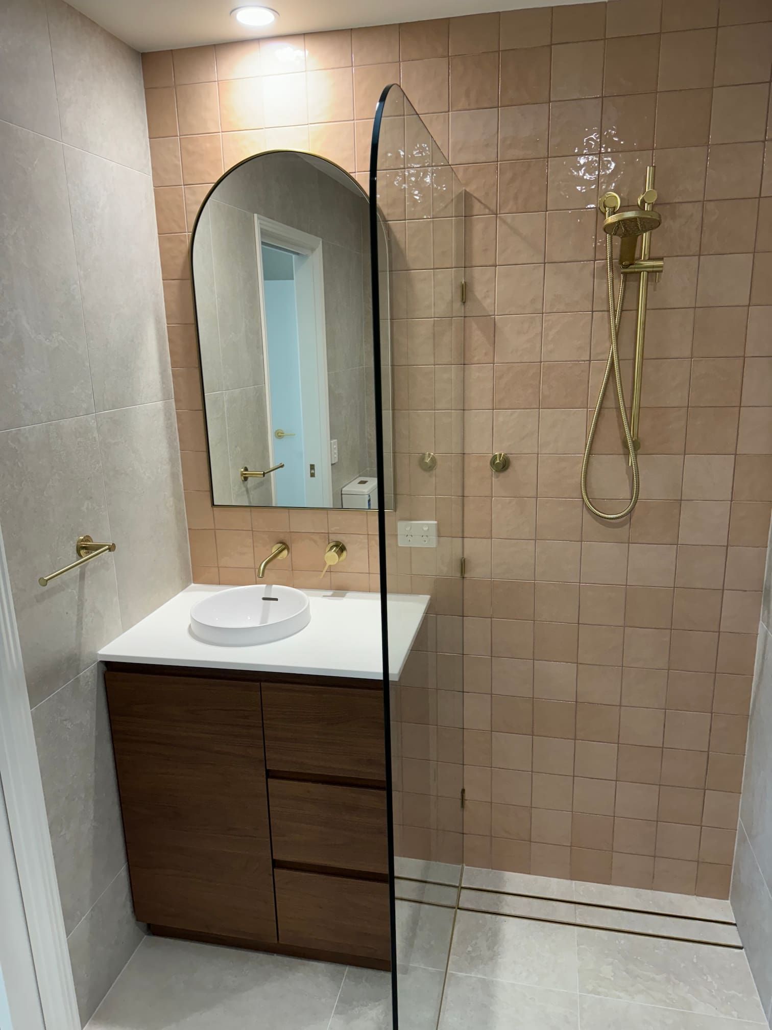 Bathroom with brown vanity, pink tiled shower, gold fixtures, and arched mirror.