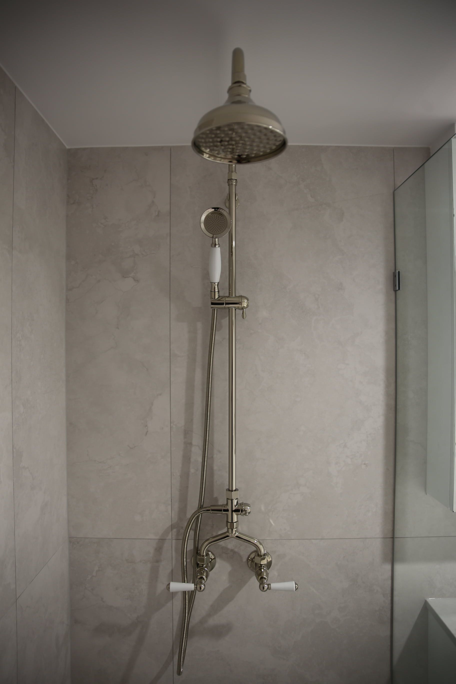 Shower fixture with overhead rain showerhead, hand-held sprayer, and vintage-style metal and porcelain fixtures on a tiled wall.
