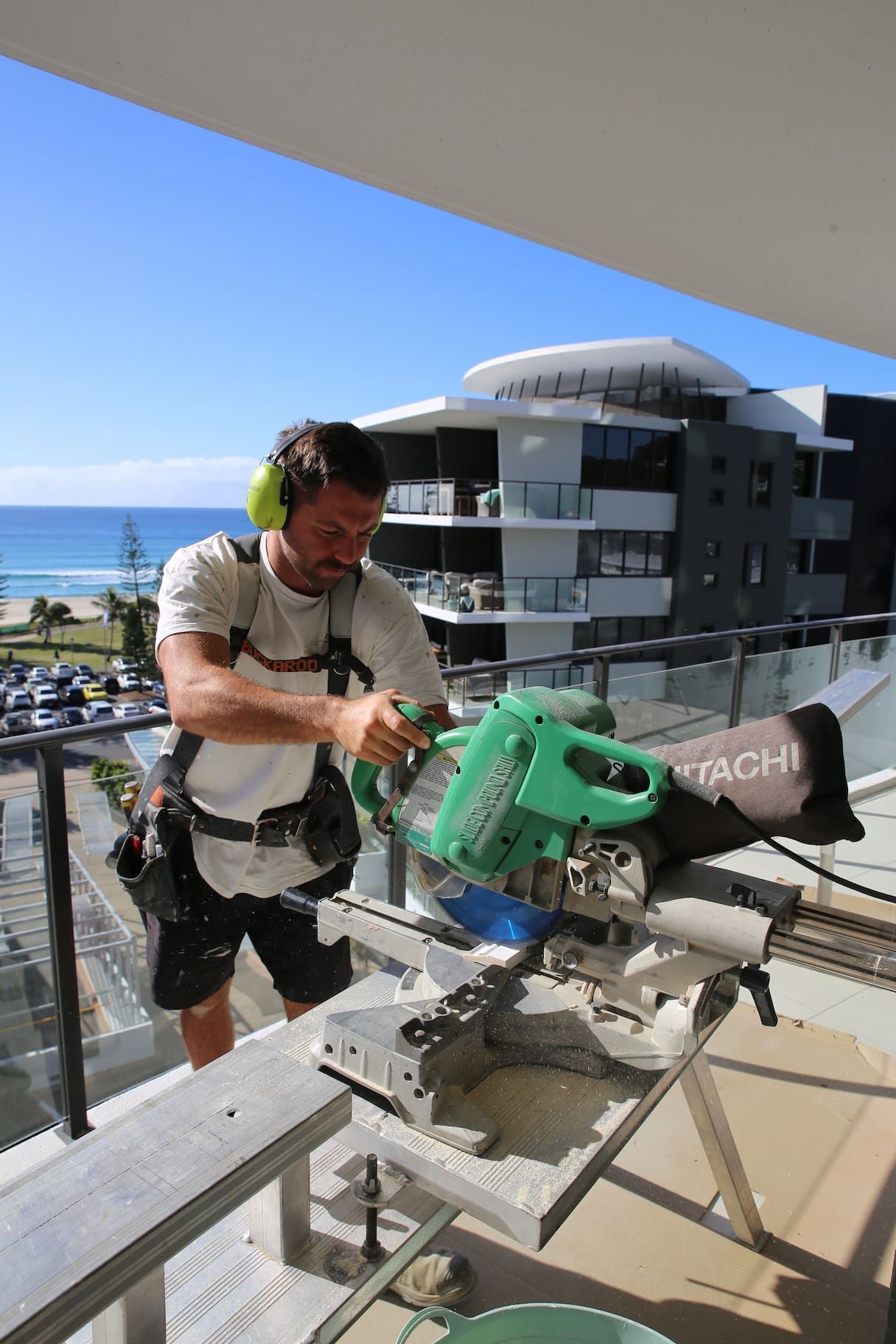 Man using a Hitachi miter saw on a balcony, ocean in background.