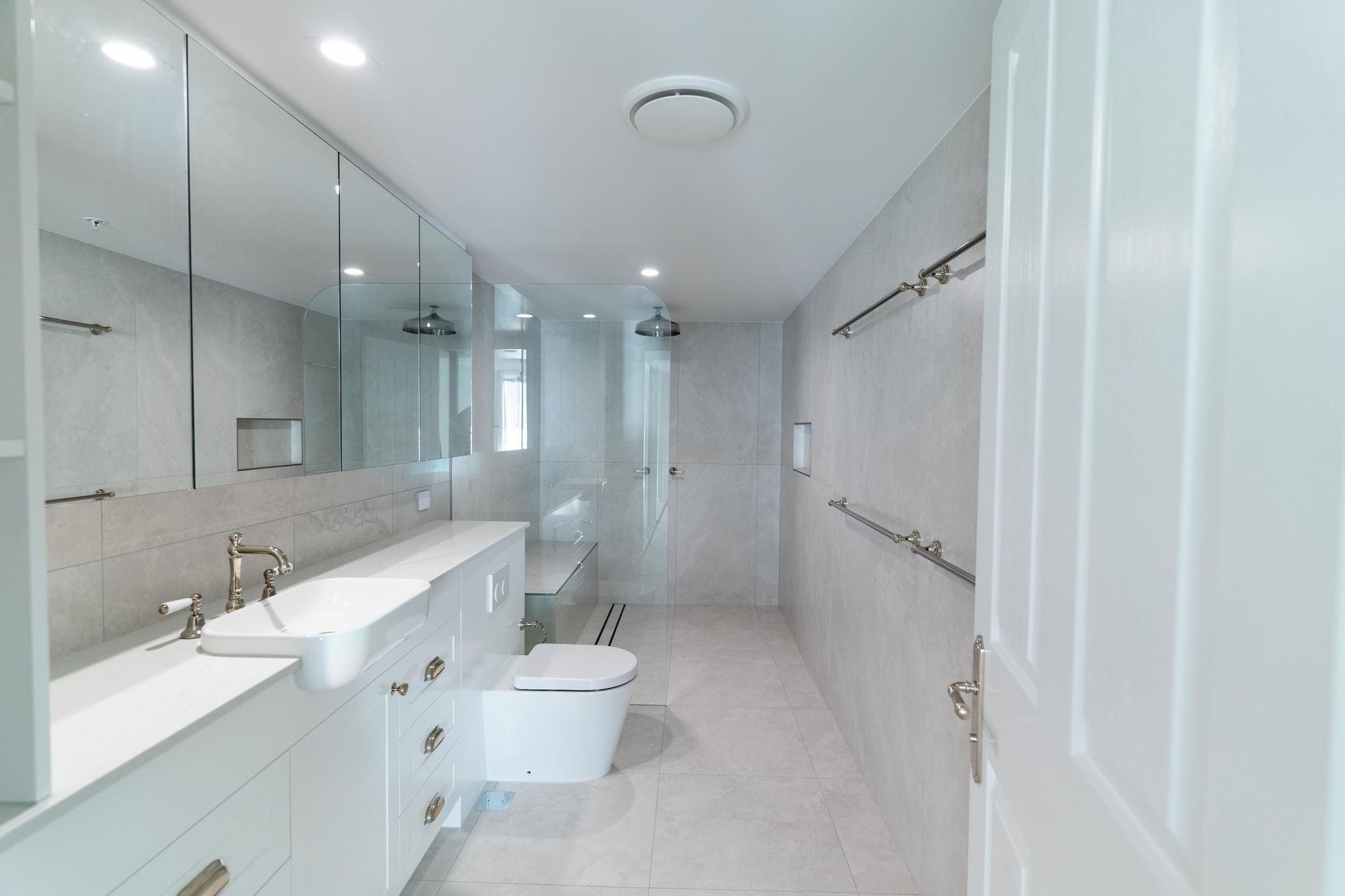 Spacious, bright white bathroom with a long vanity, toilet, and shower. Mirrors and recessed lighting.