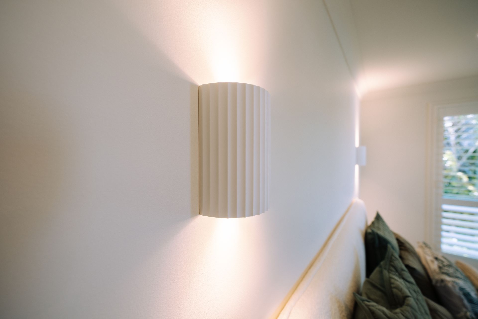 White, ribbed wall sconce illuminating a white wall, above a bed with a green quilted pillow.