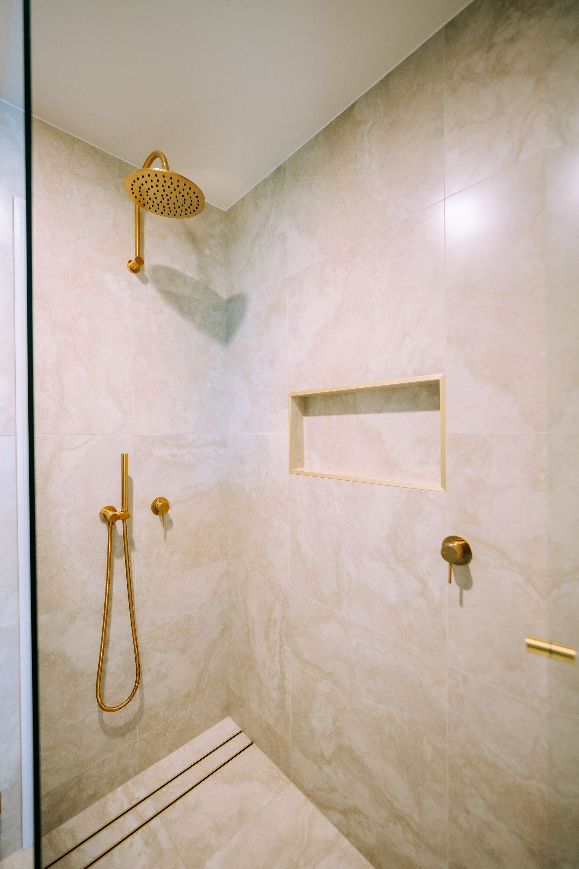 Gold shower fixtures on a light gray tile shower with a recessed shelf.
