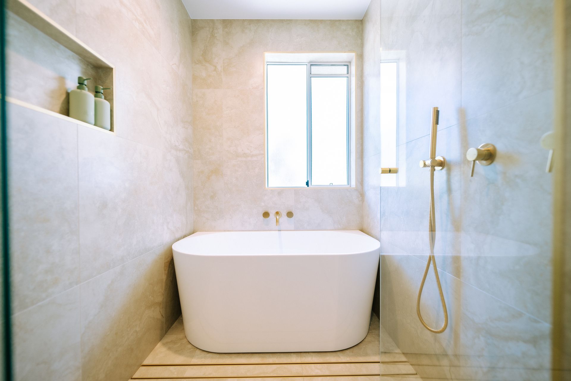 Modern bathroom with white tub, gold fixtures, and light-colored tile walls.