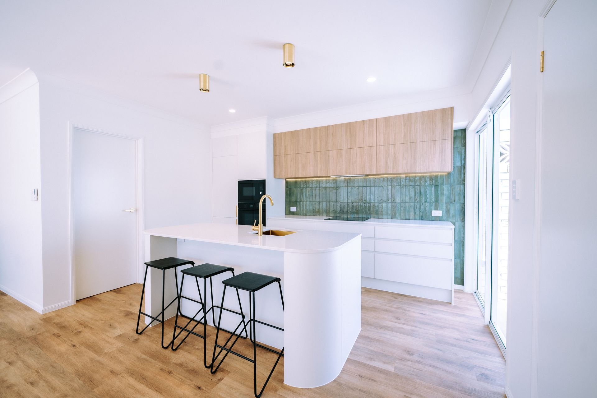 Modern kitchen with white island, black stools, light wood cabinets, and green backsplash.