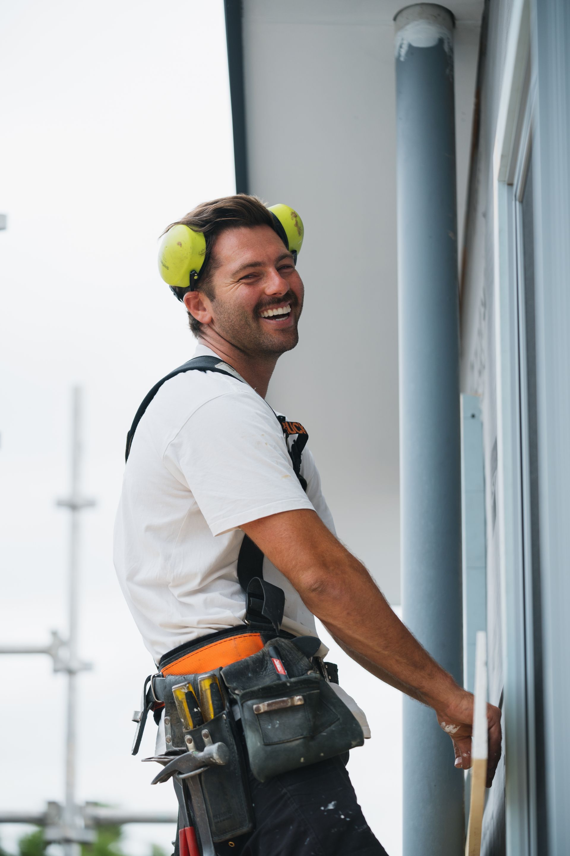 Man in work gear smiles while working on the exterior of a building, wearing ear protection.