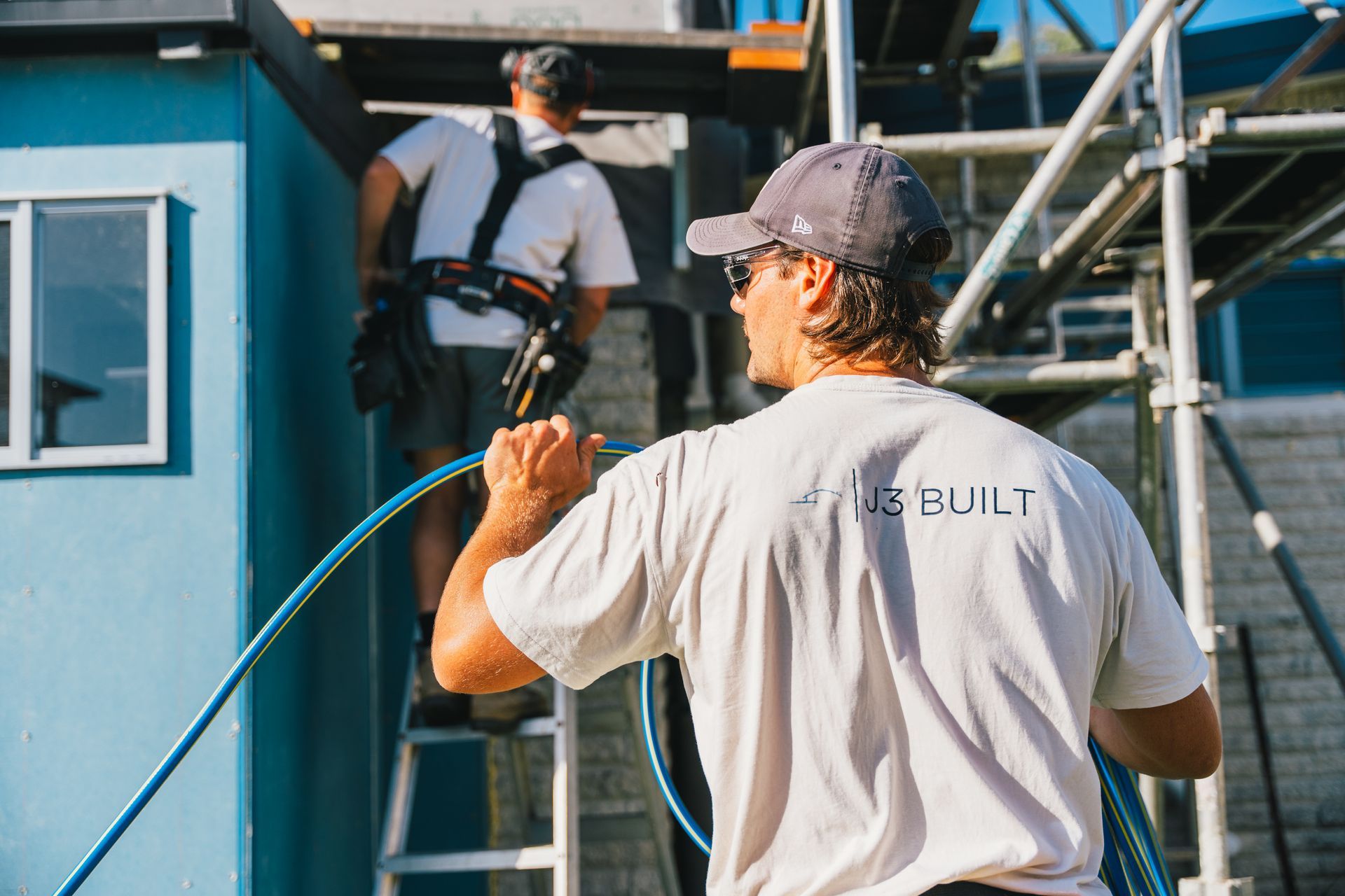 Two construction workers on a building site. One holds a blue hose, the other climbs stairs. Scaffolding is visible.