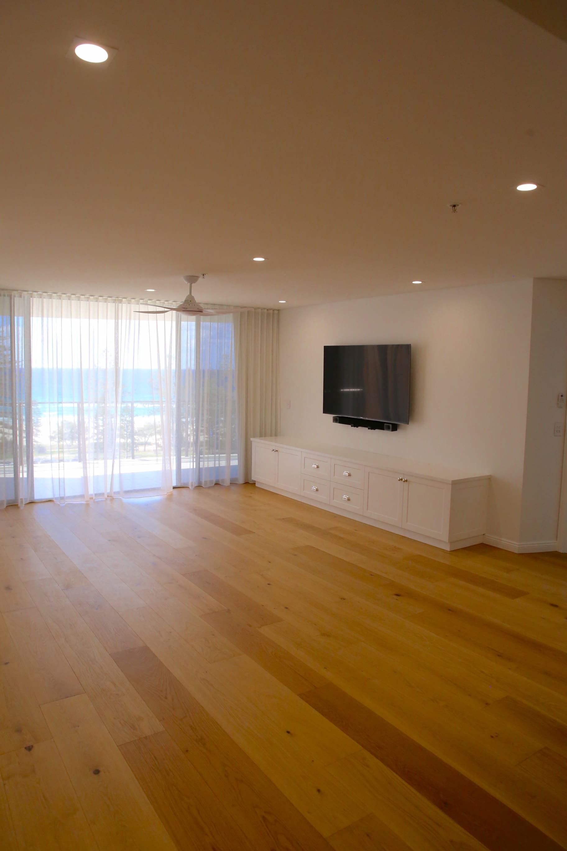 Empty living room with wood floor, white walls, TV, and large windows with sheer curtains.