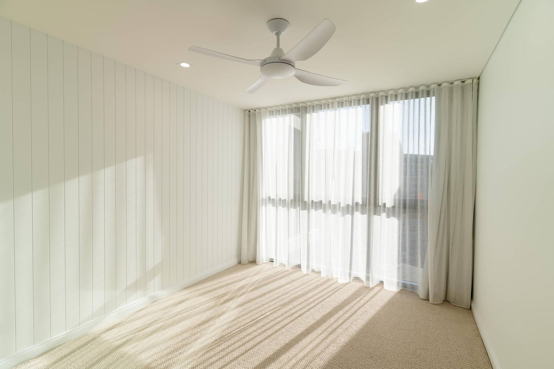 White room with a textured carpet, vertical panel wall, sheer curtains, and a ceiling fan.