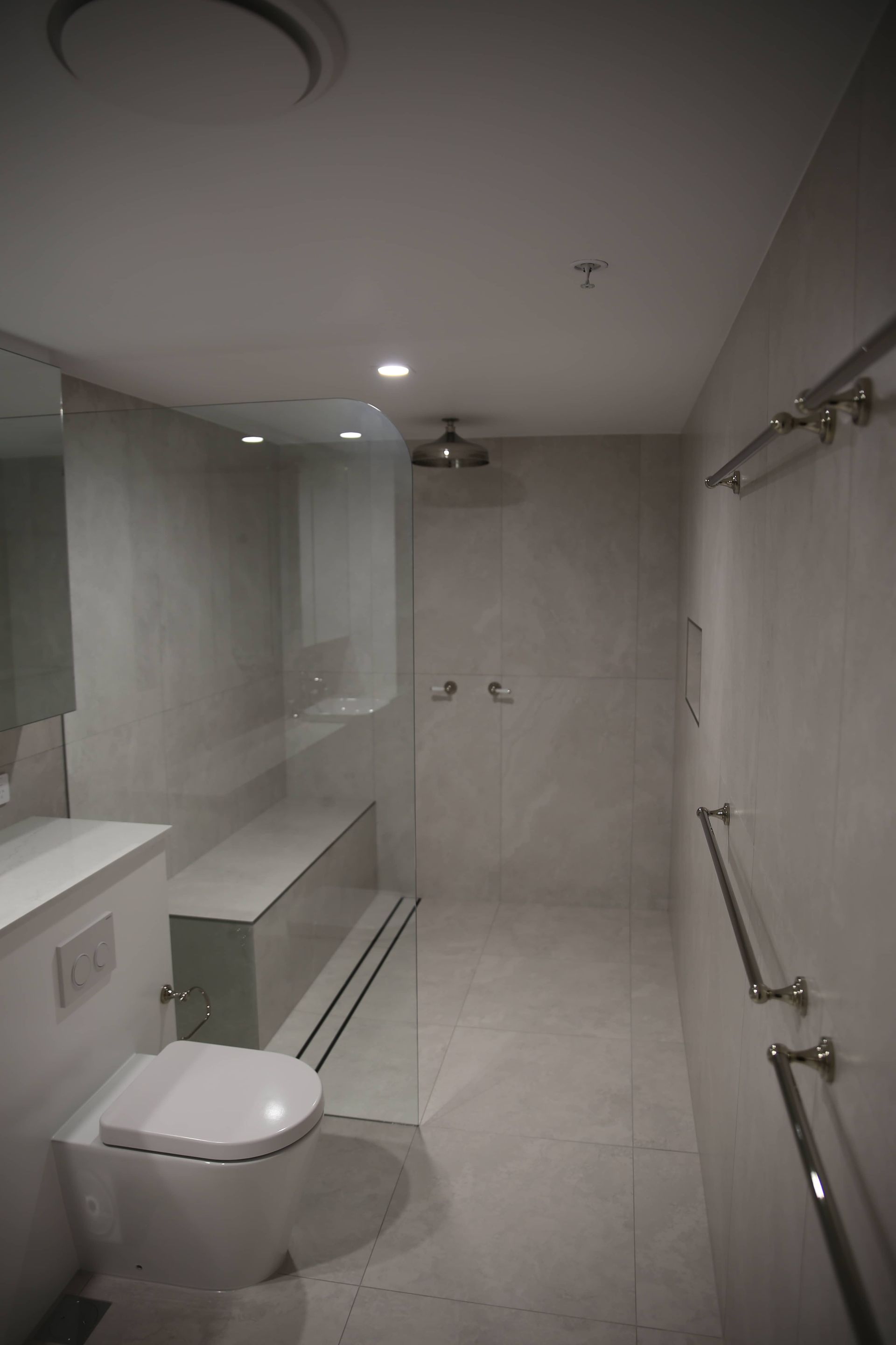 Modern, accessible bathroom with toilet, shower, grab bars, and bench. Light gray tiles and walls.