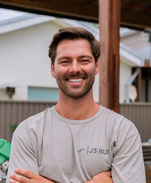 Man with brown hair and a mustache smiling, wearing a light gray shirt with a logo, standing outdoors.