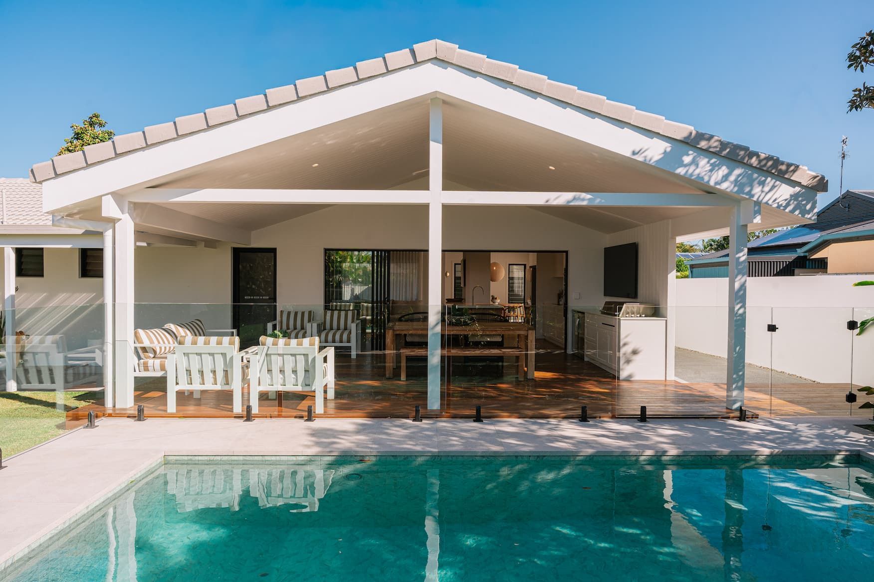 Poolside patio with pool. White structure with outdoor seating, dining table, and BBQ. Glass pool fence, blue water. Sunny day.