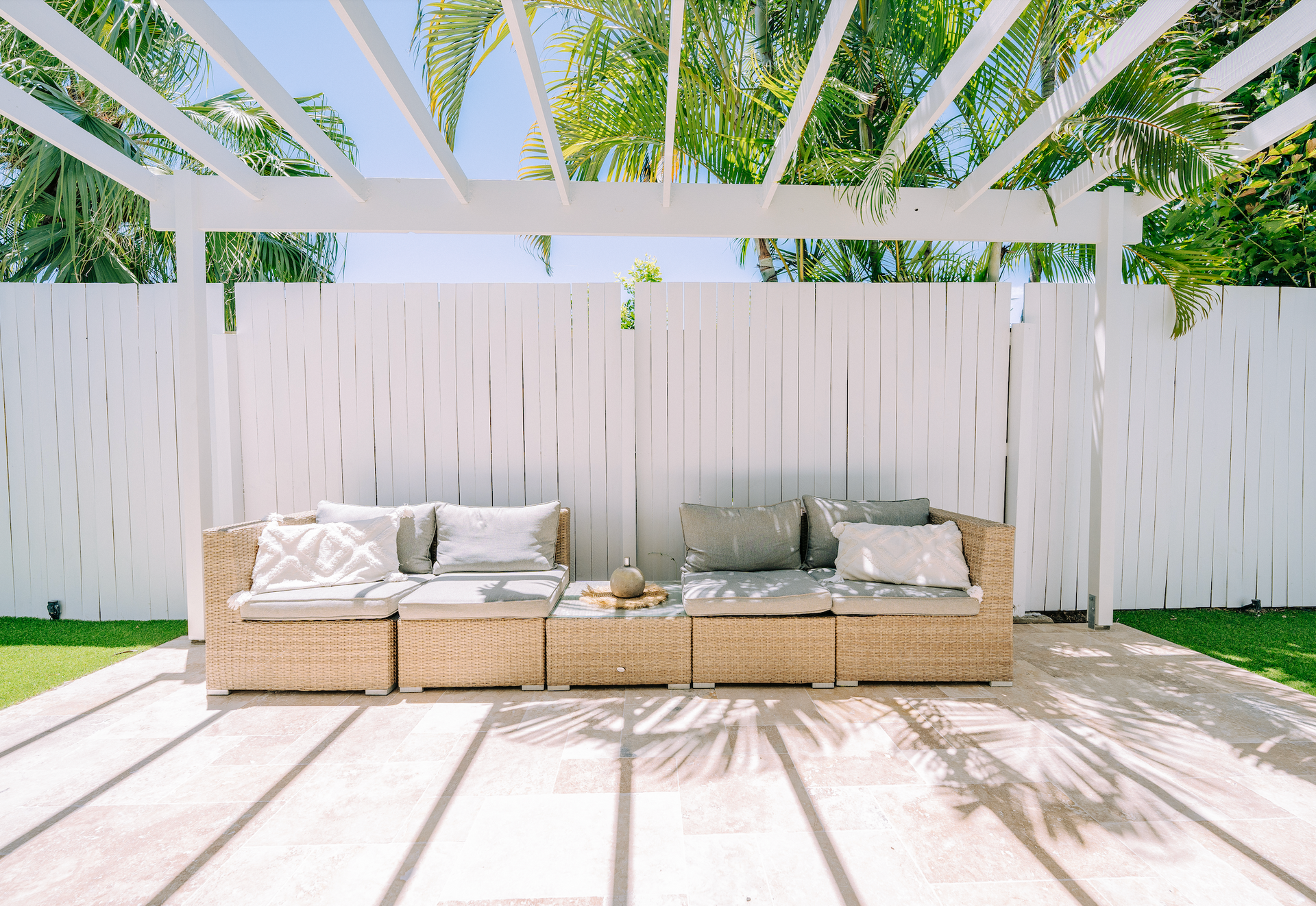 Outdoor seating area with white pergola, beige wicker furniture, and white wooden fence.