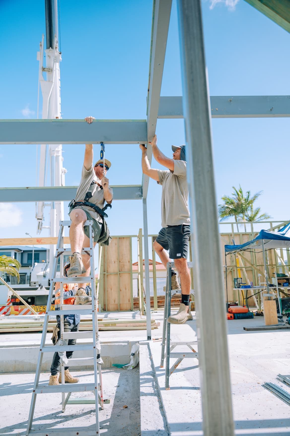 Two construction workers on ladders installing metal beams at a building site under a blue sky.