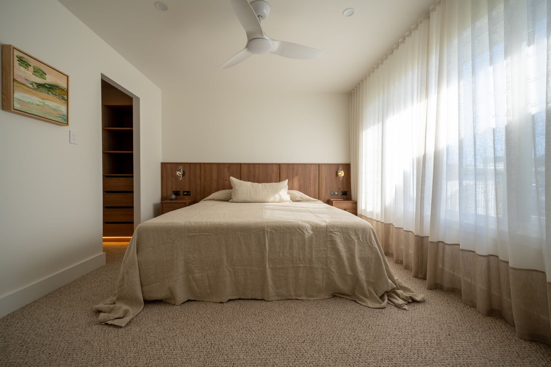 Bedroom with beige bedding, wooden headboard, walk-in closet, and sheer curtains.