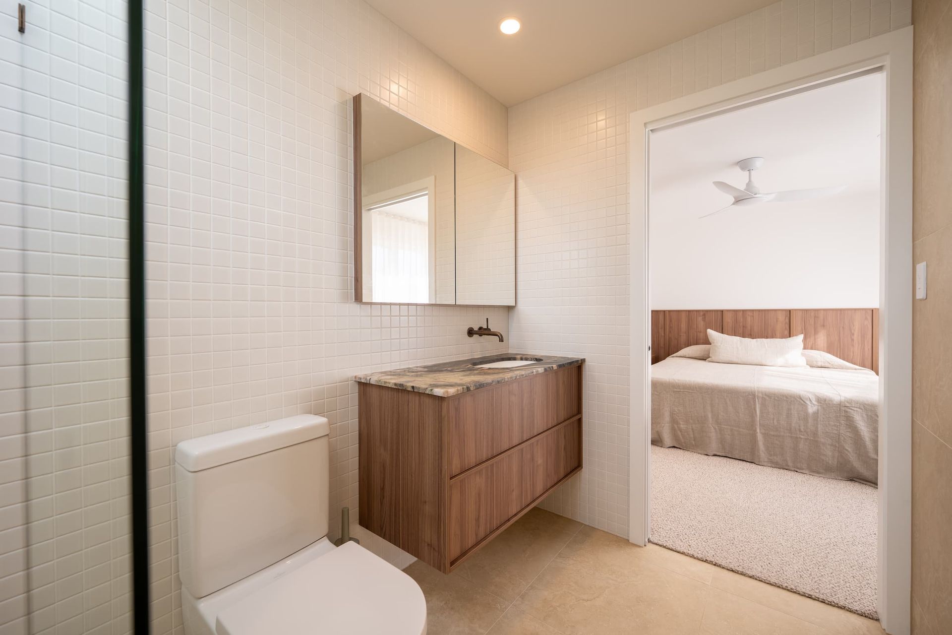 Bathroom with white tile walls, wood vanity, and open doorway to a bedroom with a bed.
