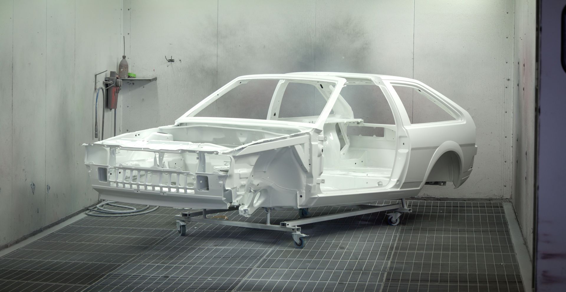 White car body shell in a paint booth, mounted on a stand during restoration or fabrication.