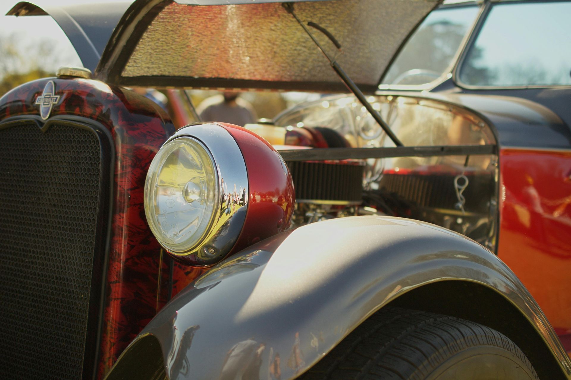 Close-up of a vintage red car with chrome headlight and gray fender under an open hood.