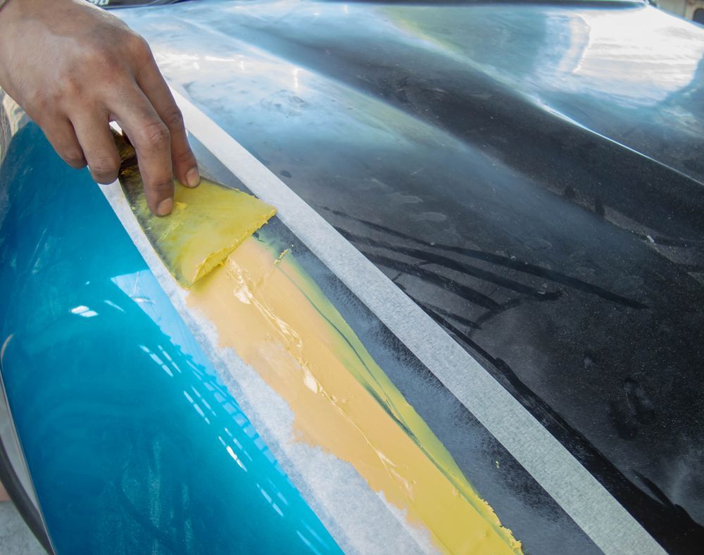 Hand sanding yellow filler on a blue car panel beside masking tape on a windshield