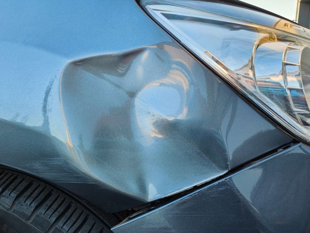 Close-up of a blue car’s front fender with a large dent near the headlight.