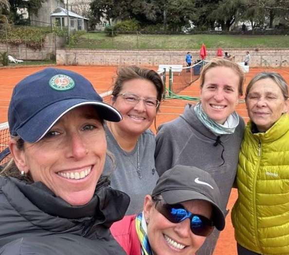 Cinco personas sonrientes en una cancha de tenis de arcilla roja, vestidas con ropa informal, posando para una selfie.