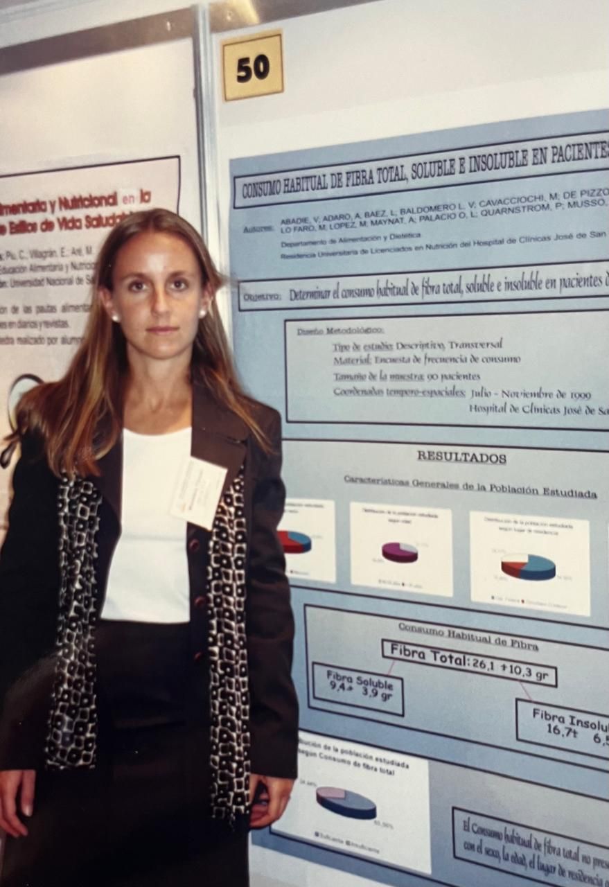 Mujer presentando un poster de investigación, vestida con traje, junto a una vitrina numerada.