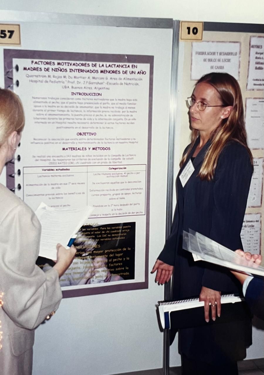 Mujer presentando un póster científico en una conferencia. Sostiene artículos y gesticula, explicando la investigación al público.
