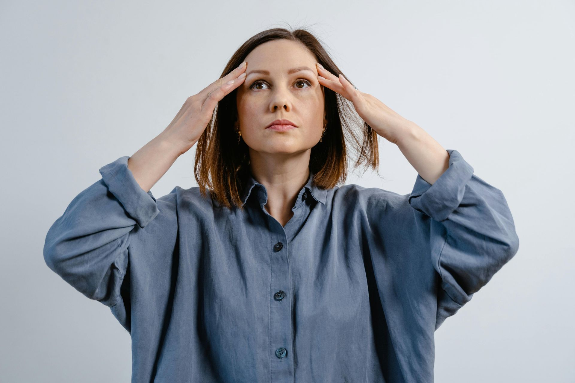 A person in a blue button-down shirt holding their head with both hands, looking upward with a pained expression.