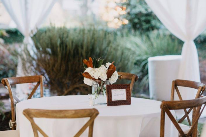 Round outdoor table with wooden chairs and a white floral centerpiece under a white canopy.