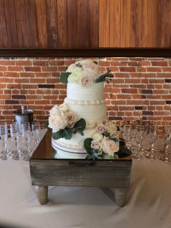 Three-tier white wedding cake with flowers on a rustic table, flanked by glassware, against a brick wall
