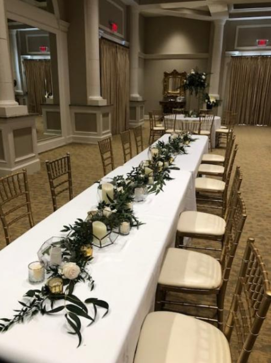 Long banquet table with white tablecloth, gold chairs, and green floral garland in an elegant hall