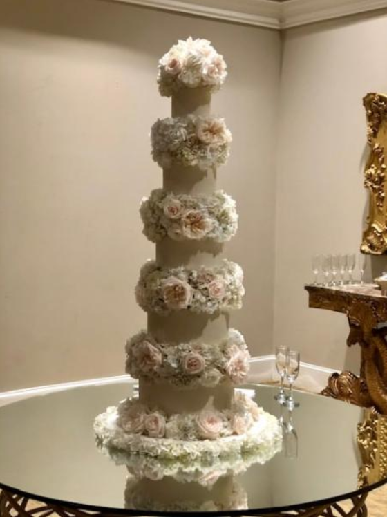 Tall multi-tier white floral wedding cake on a mirrored table in a formal room