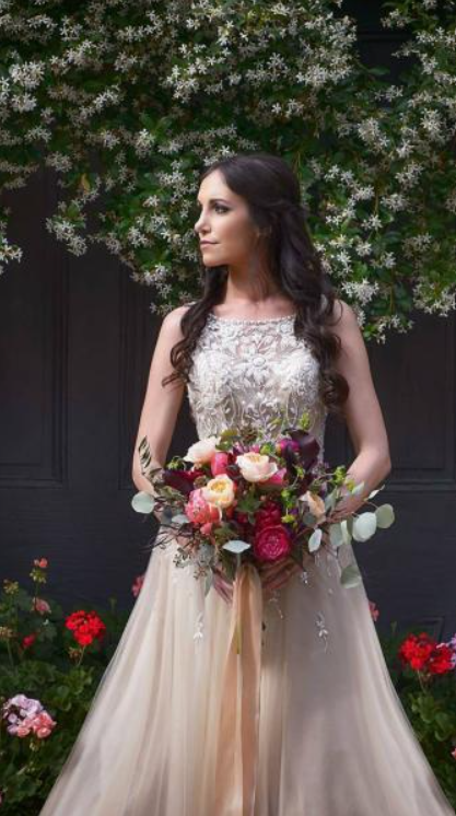 Woman in a cream gown holding a colorful bouquet, standing outdoors before dark doors and blooming vines