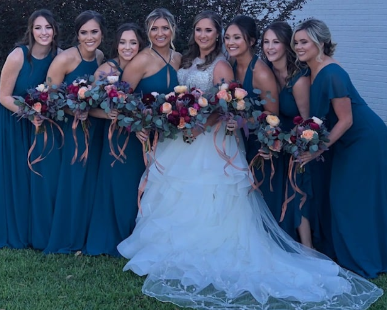 Bride in white gown with six women in navy dresses holding floral bouquets outdoors at dusk