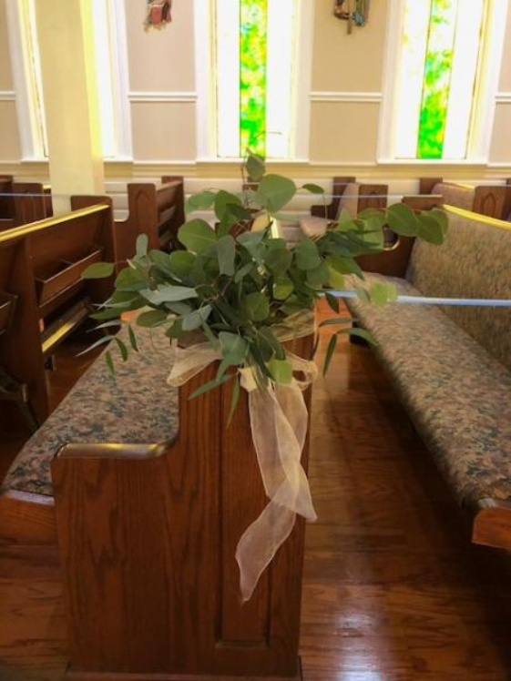 Bouquet of greenery with white ribbon tied to a church pew in a sunlit chapel