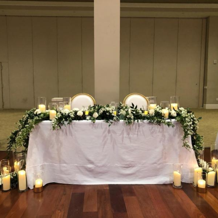 Elegant banquet table draped in white, decorated with greenery, candles, and gold-rimmed plates in a formal hall