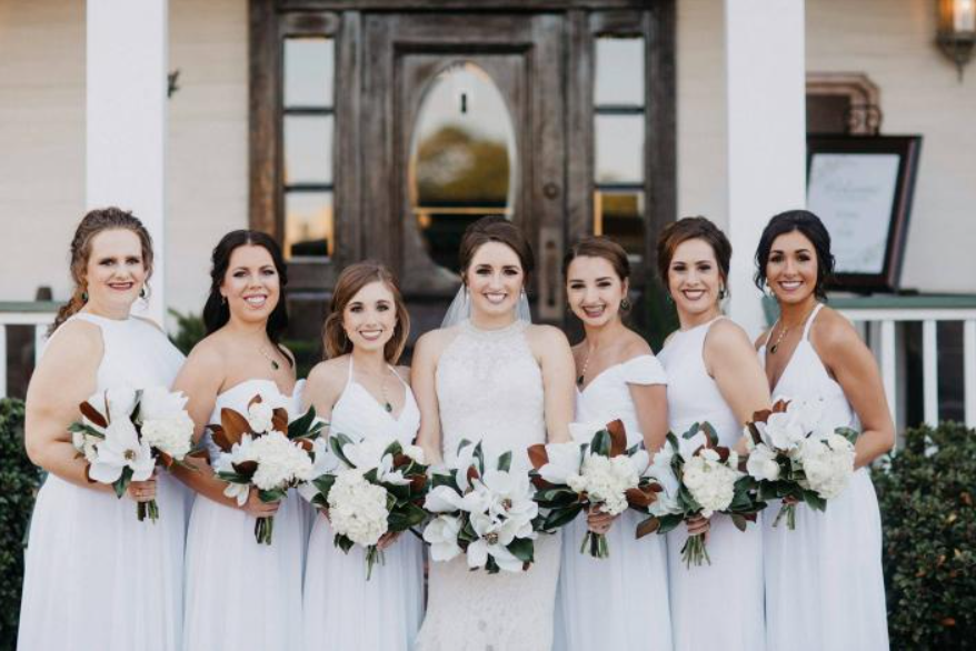 Six bridesmaids in white dresses holding bouquets in front of a building entrance