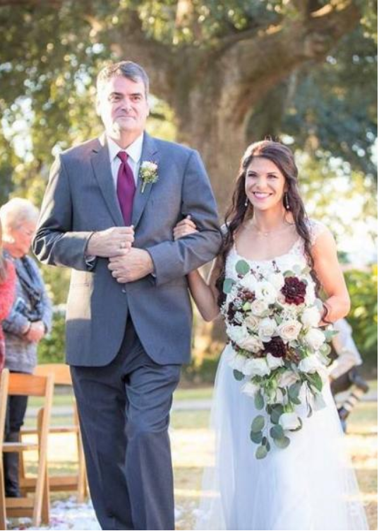 Bride in white lace dress walks arm-in-arm with man in gray suit, holding bouquet outdoors at a wedding