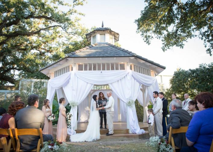 Wedding ceremony under white drapes at a gazebo, with bride and groom standing at the altar outdoors.