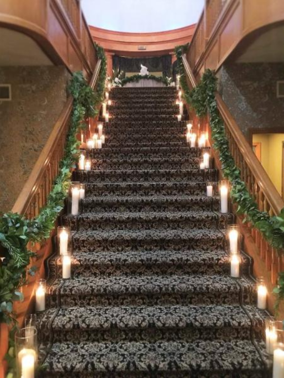 Grand staircase with patterned carpet, candle-like lights, and green plants in a grand interior lobby