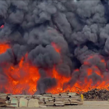 Large fire with thick black smoke billowing over stacked materials in an industrial area