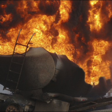 A large fire blazes behind damaged industrial tanks and metal equipment.