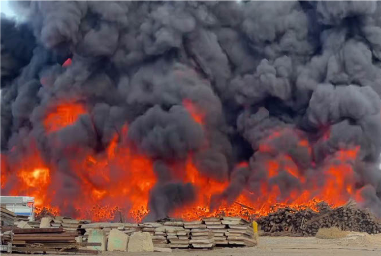 Massive blaze with thick black smoke engulfing a scrap yard, orange flames behind stacked materials