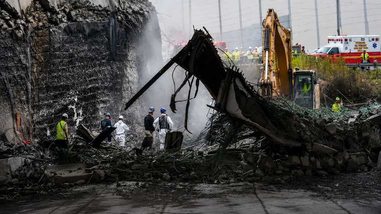 Collapsed dark structure beside water with people and a yellow excavator at the site