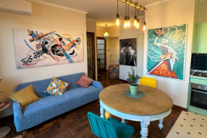 Soggiorno con opere d'arte astratte, divano blu, tavolo rotondo, sedie colorate e pavimento in legno.