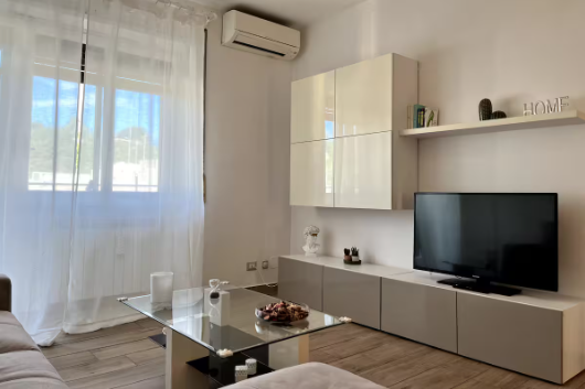 Soggiorno con TV, armadi, tavolo in vetro e una finestra con tende bianche.
